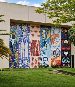 Ateliê Udo Knoff e 12 artistas transformam a Reitoria da Uefs em galeria a céu aberto Dois grandes murais de azulejos integram arte e arquitetura no campus e valorizam diferentes expressões da cultura nordestina Nas paredes laterais da Reitoria da Universidade Estadual de Feira de Santana (Uefs), a arte participa do cotidiano de quem estuda, trabalha ou visita o campus. Datado de 2002, o conjunto formado por dois grandes murais de azulejos transforma o prédio em uma galeria a céu aberto, com figuras, símbolos e composições que convidam à observação e aproximam a comunidade universitária da produção artística baiana. São 12 painéis, seis de cada lado, com obras de Antônio Brasileiro, Bel Borba, Juraci Dórea, Justino Marinho, Washington Falcão, Juarez Paraíso, Chico Liberato, Reinaldo Eckenberger, Sônia Rangel, César Romero, J. Cunha e Márcia Magno. A variedade de linguagens permite reconhecer diferentes trajetórias e maneiras de interpretar a cultura, compondo um conjunto de forte presença na paisagem universitária. Uma escolha que valoriza a diversidade A iniciativa merece reconhecimento tanto pela dimensão monumental dos murais quanto pela decisão de prestigiar diversos artistas consagrados. Cada autor recebeu uma ampla superfície para desenvolver uma criação própria. A reunião dessas obras valoriza a pluralidade da arte baiana e demonstra como diferentes estilos podem compartilhar um espaço sem perder a identidade. Os murais estão ligados ao Projeto Nordeste de Artes Plásticas, movimento que promoveu exposições, encontros e intercâmbios na região durante a década de 1980. Na Uefs, esse espírito coletivo ganhou permanência na arquitetura. O conjunto amplia a experiência de estar no campus e reafirma a importância da arte na formação cultural, no exercício da sensibilidade e na construção da memória. O legado do Ateliê Udo Knoff Os azulejos foram confeccionados no Ateliê Cerâmico Udo Knoff, em Salvador. Fundado pelo ceramista alemão radicado na Bahia e instalado no bairro de Brotas, o espaço tornou-se referência na integração entre arte e arquitetura. Knoff executou composições de diferentes artistas e incentivou a exploração da cerâmica como linguagem. A transferência dos desenhos, a pintura e a queima das peças reuniam criação artística, trabalho artesanal e conhecimento técnico. A permanência desse patrimônio exige cuidado contínuo. Em visita ao local, foi possível observar perda de brilho em parte dos azulejos do lado oeste do prédio, que recebe o sol da tarde. A observação reforça a importância de uma avaliação especializada para identificar as causas e orientar a conservação. Preservar os murais é também proteger a memória dos artistas e uma iniciativa que deu à arte lugar de destaque na universidade. Everaldo Goes | © Feira Hoje Siga @feirahoje 11/10/26