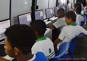 Computação na rede municipal de Feira vai além de aprender a usar o computador Diretrizes preveem raciocínio lógico, compreensão das tecnologias e uso responsável da internet, com implantação gradual e conteúdos integrados ao currículo Resolver problemas, compreender como as tecnologias funcionam e aprender a avaliar as informações que circulam na internet. Esses são alguns dos objetivos do ensino de Computação na Educação Básica, que ganhou um novo encaminhamento na rede municipal de Feira de Santana. Em notícia publicada em 13 de outubro, a Prefeitura informou a adesão às diretrizes curriculares da Bahia, formalizada pelo Conselho Municipal de Educação por meio da Resolução nº 030/2026. A implantação será gradual, acompanhada de formação dos profissionais e incorporada ao planejamento das escolas. A proposta organiza a aprendizagem em três eixos. O pensamento computacional desenvolve a capacidade de dividir problemas em partes, identificar padrões e estabelecer passos para encontrar soluções. O mundo digital envolve a compreensão de equipamentos, redes e do armazenamento e da circulação de dados. Já a cultura digital trata da participação consciente, crítica e responsável nos ambientes tecnológicos. Na prática, a formação busca dar aos estudantes condições de compreender e utilizar a tecnologia com maior autonomia. Aprendizagem também sem telas Na educação infantil, essa abordagem pode envolver brincadeiras de organizar objetos, reconhecer repetições e seguir sequências de instruções. São possibilidades da chamada computação desplugada, realizada sem computador. A Resolução CME nº 90/2025, publicada em agosto do ano passado, já previa atividades lúdicas e exploração de padrões nessa etapa. Para o ensino fundamental, estabelecia a introdução progressiva de programação, robótica e projetos interdisciplinares. Na Educação de Jovens e Adultos, a orientação é trabalhar situações do cotidiano e tecnologias acessíveis. O mesmo documento municipal define a Computação como componente transversal e integrador, articulado às demais áreas do currículo. Assim, uma atividade de Matemática pode explorar sequências e soluções para problemas, enquanto uma proposta de leitura pode estimular a avaliação das informações recebidas. Esses são exemplos possíveis de aplicação. A norma também atribui à Secretaria Municipal de Educação a responsabilidade de assegurar infraestrutura tecnológica mínima, materiais pedagógicos e formação continuada dos professores. Feira integra orientação nacional A mudança faz parte de um movimento mais amplo. Em 2022, o Conselho Nacional de Educação estabeleceu as normas de Computação como complemento à Base Nacional Comum Curricular. As diretrizes nacionais de educação digital e midiática de 2025 fixaram o início da implementação dos novos currículos em 2026. Na Bahia, a regulamentação estadual admite a adesão dos sistemas municipais, respeitando a autonomia de cada rede. A decisão de Feira insere-se nesse processo, com a expectativa de desenvolver raciocínio lógico, criatividade, segurança digital e capacidade de analisar informações. A rede em Feira de Santana já registra ações relacionadas ao tema. Em março deste ano, uma formação dos profissionais dos anos iniciais apresentou o projeto MicroKids, associado pela Seduc às diretrizes da BNCC Computação. Em abril, professores receberam orientações sobre a plataforma e o uso de recursos tecnológicos em diferentes componentes curriculares. A resolução de 2026 determina que as escolas incorporem as competências e habilidades aos projetos pedagógicos e prevê acompanhamento pela Secretaria de Educação e pelo Conselho Municipal. O documento não estabelece datas de implantação por escola, valores de investimento ou quantitativos de equipamentos. Computação na Educação Básica, educação digital, Feira de Santana, rede municipal de ensino, BNCC Computação, pensamento computacional, cultura digital, formação de professores, Conselho Municipal de Educação, Secretaria Municipal de Educação Por Everaldo Goes, com informações Ministério da Educação, Secretaria de Educação do Estado e Secretaria Municipal de Educação Siga @feirahoje 15/10/26