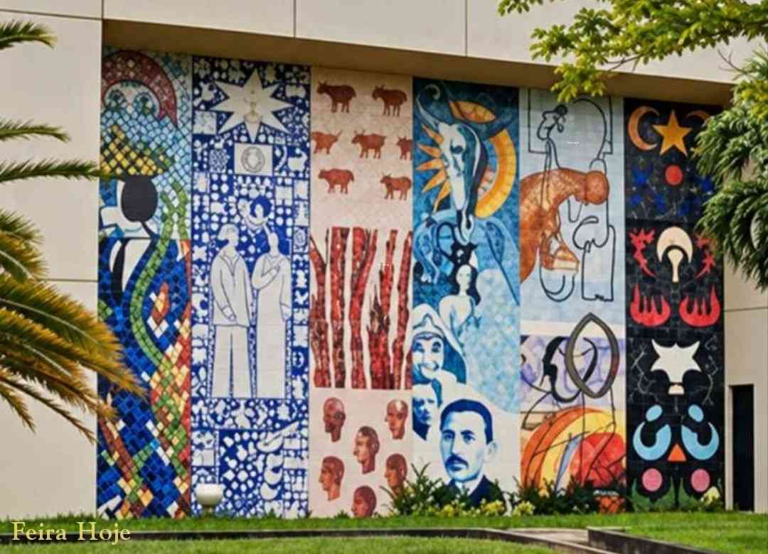 Ateliê Udo Knoff e 12 artistas transformam a Reitoria da Uefs em galeria a céu aberto Dois grandes murais de azulejos integram arte e arquitetura no campus e valorizam diferentes expressões da cultura nordestina Nas paredes laterais da Reitoria da Universidade Estadual de Feira de Santana (Uefs), a arte participa do cotidiano de quem estuda, trabalha ou visita o campus. Datado de 2002, o conjunto formado por dois grandes murais de azulejos transforma o prédio em uma galeria a céu aberto, com figuras, símbolos e composições que convidam à observação e aproximam a comunidade universitária da produção artística baiana. São 12 painéis, seis de cada lado, com obras de Antônio Brasileiro, Bel Borba, Juraci Dórea, Justino Marinho, Washington Falcão, Juarez Paraíso, Chico Liberato, Reinaldo Eckenberger, Sônia Rangel, César Romero, J. Cunha e Márcia Magno. A variedade de linguagens permite reconhecer diferentes trajetórias e maneiras de interpretar a cultura, compondo um conjunto de forte presença na paisagem universitária. Uma escolha que valoriza a diversidade A iniciativa merece reconhecimento tanto pela dimensão monumental dos murais quanto pela decisão de prestigiar diversos artistas consagrados. Cada autor recebeu uma ampla superfície para desenvolver uma criação própria. A reunião dessas obras valoriza a pluralidade da arte baiana e demonstra como diferentes estilos podem compartilhar um espaço sem perder a identidade. Os murais estão ligados ao Projeto Nordeste de Artes Plásticas, movimento que promoveu exposições, encontros e intercâmbios na região durante a década de 1980. Na Uefs, esse espírito coletivo ganhou permanência na arquitetura. O conjunto amplia a experiência de estar no campus e reafirma a importância da arte na formação cultural, no exercício da sensibilidade e na construção da memória. O legado do Ateliê Udo Knoff Os azulejos foram confeccionados no Ateliê Cerâmico Udo Knoff, em Salvador. Fundado pelo ceramista alemão radicado na Bahia e instalado no bairro de Brotas, o espaço tornou-se referência na integração entre arte e arquitetura. Knoff executou composições de diferentes artistas e incentivou a exploração da cerâmica como linguagem. A transferência dos desenhos, a pintura e a queima das peças reuniam criação artística, trabalho artesanal e conhecimento técnico. A permanência desse patrimônio exige cuidado contínuo. Em visita ao local, foi possível observar perda de brilho em parte dos azulejos do lado oeste do prédio, que recebe o sol da tarde. A observação reforça a importância de uma avaliação especializada para identificar as causas e orientar a conservação. Preservar os murais é também proteger a memória dos artistas e uma iniciativa que deu à arte lugar de destaque na universidade. Everaldo Goes | © Feira Hoje Siga @feirahoje 11/10/26